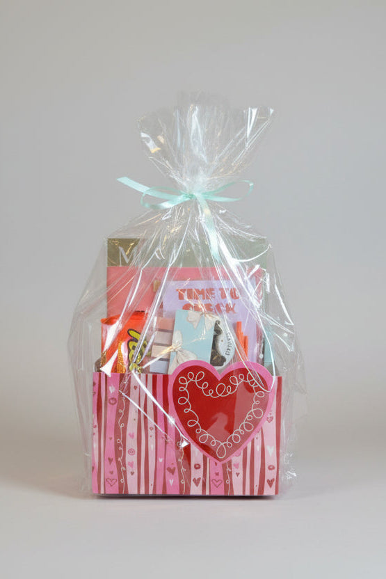 Gift basket with a pink bag and heart design, tied with cellophane and a bow, on a wooden surface.